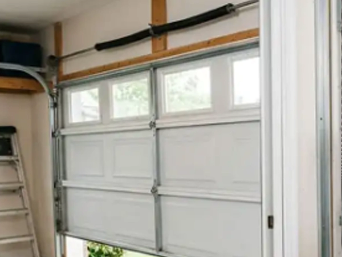 Inside a residential garage showing a white sectional door with small window panels at the top; a ladder leans against the wall to the left.